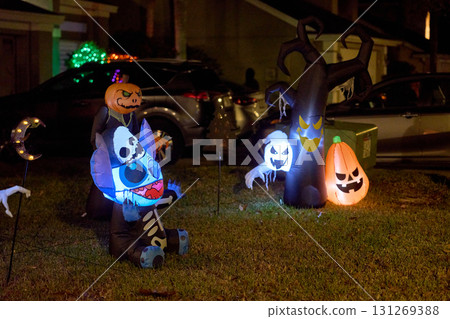 Blue alien skeleton, ghost, pumpkin, and spooky tree inflatables glow on a suburban lawn at night. Halloween yard decorations with bright lights create a festive, eerie holiday display. 131269388