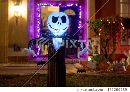 A tall inflatable scarecrow with a glowing skeleton head stands in a suburban front yard at night. Halloween lights, bats, and decorations create a spooky and festive scene. 131269389