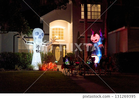 Big inflatable skeleton and monster glow outside a welcoming house at night. Neighbors sit with candy ready for trick-or-treaters. Big inflatable skeleton and monster glow outside a welcoming house at night. Neighbors sit with candy ready for trick-or-treaters. 131269395