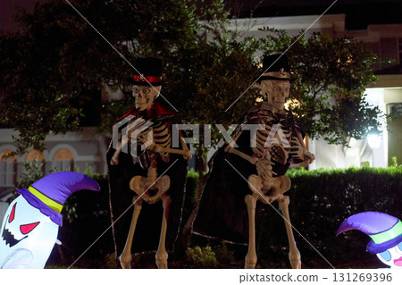 Two skeletons in top hats and capes pose with instruments on the grass. Spooky night yard display with glowing inflatables nearby. 131269396