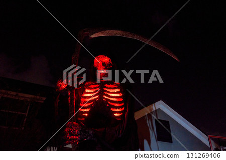 Hooded skeleton reaper glows red with bright ribs and eyes beside a house. Dramatic Halloween yard display with large scythe and deep shadows. Hooded skeleton reaper glows red with bright ribs and eyes beside a house. Dramatic Halloween yard display with large scythe and deep shadows. 131269406