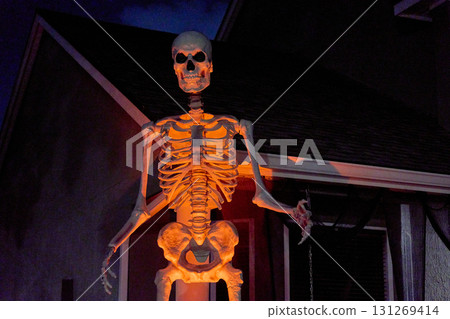 A towering skeleton figure stands by a house porch under orange spotlight. Spooky Halloween yard prop with bones and skull dominates the dark scene. A towering skeleton figure stands by a house porch under orange spotlight. Spooky Halloween yard prop with bones and skull dominates the dark scene. 131269414