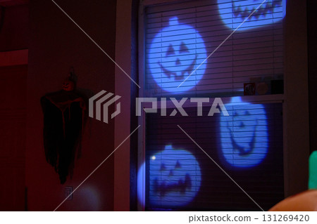 Several smiling jack-o-lantern faces are projected in blue light on a home window. Festive Halloween display with spooky yet cheerful mood at night. 131269420
