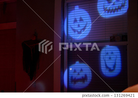 Smiling pumpkin faces are projected in blue light onto a home window at night. Festive Halloween decoration with a spooky yet cheerful mood. 131269421