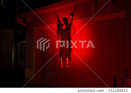 A hooded reaper with bony legs hangs on a garage door under deep red lighting. Spooky Halloween yard prop with dramatic shadows at night. A hooded reaper with bony legs hangs on a garage door under deep red lighting. Spooky Halloween yard prop with dramatic shadows at night. 131269422