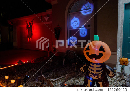 Front yard with a smiling inflatable pumpkin, blue jack-o-lantern window projections, and a red-lit reaper. Festive Halloween home display at night with spooky lights. Front yard with a smiling inflatable pumpkin, blue jack-o-lantern window projections, and a red-lit reaper. Festive Halloween home display at night with spooky lights. 131269423