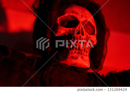 Close-up skull in a hooded cloak under deep red light at night. Spooky Halloween decoration with dramatic shadows and texture. 131269429