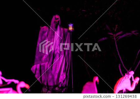 Two tall hooded reapers with lanterns glow purple in a spooky lawn scene. The nighttime haunted house yard is filled with eerie lights and shadows. Two tall hooded reapers with lanterns glow purple in a spooky lawn scene. The nighttime haunted house yard is filled with eerie lights and shadows. 131269438
