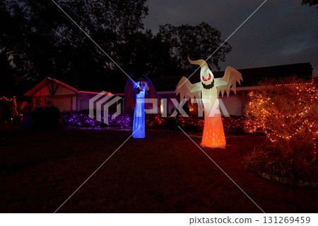 Front yard house displays a glowing inflatable ghost and a tall blue winged demon at night. Colorful lights and bushes create a spooky Halloween display. 131269459