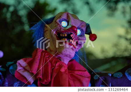 A sinister clown stares with bright spiral eyes and a red nose under night light. Blue hair, rough wrinkles, and big ruffles make a spooky Halloween display. A sinister clown stares with bright spiral eyes and a red nose under night light. Blue hair, rough wrinkles, and big ruffles make a spooky Halloween display. 131269464