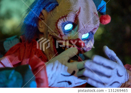 Closeup of a creepy clown with blue glowing spiral eyes and a red nose. White gloves, yellow teeth, and dark night lighting create a scary Halloween mood. Closeup of a creepy clown with blue glowing spiral eyes and a red nose. White gloves, yellow teeth, and dark night lighting create a scary Halloween mood. 131269465