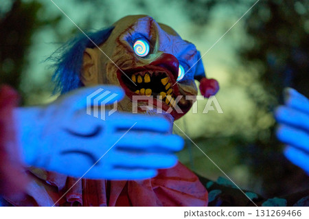 Creepy clown reaches forward with blue-lit hands and hypnotic spiral eyes. Red nose, yellow teeth, and dramatic lighting create a scary Halloween scene. Creepy clown reaches forward with blue-lit hands and hypnotic spiral eyes. Red nose, yellow teeth, and dramatic lighting create a scary Halloween scene. 131269466