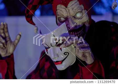 Creepy jester in red and black holds a white clown mask under blue night light. Glowing eyes, sharp teeth, and dramatic costume make a spooky Halloween scene. Creepy jester in red and black holds a white clown mask under blue night light. Glowing eyes, sharp teeth, and dramatic costume make a spooky Halloween scene. 131269470