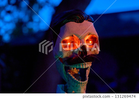 Closeup portrait of a skull with a mustache and bright red eyes against blue twilight. Spooky Halloween prop creates an eerie yard mood. 131269474