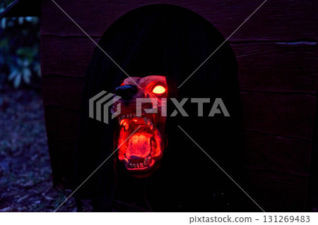Snarling monster dog head glows red from a dark doghouse doorway at night. Sharp fangs, bright eyes, and dramatic lighting create a scary Halloween yard prop. Snarling monster dog head glows red from a dark doghouse doorway at night. Sharp fangs, bright eyes, and dramatic lighting create a scary Halloween yard prop. 131269483