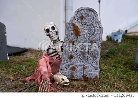 Spooky Halloween decoration shows a weathered skeleton leaning on a cracked tombstone in a front yard. Tattered cloth, fake moss, and a small animal skeleton add creepy graveyard detail. 131269485