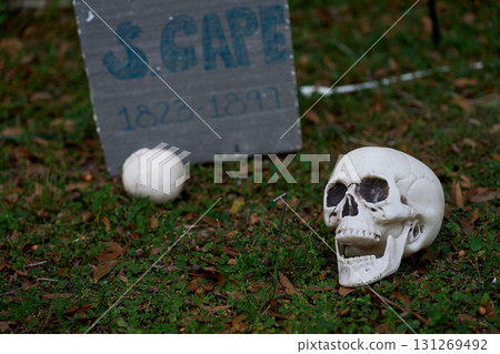 A human skull lies beside a weathered tombstone in the cemetery grass. The bony head and open jaw create a spooky Halloween scene. A human skull lies beside a weathered tombstone in the cemetery grass. The bony head and open jaw create a spooky Halloween scene. 131269492