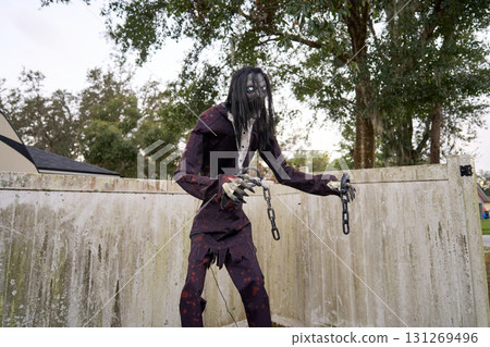 A tall haunted figure with long black hair holds hanging chains by a weathered fence. The creepy monster wears a tattered suit and stares with eerie glowing eyes. 131269496