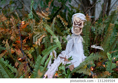 A small skeleton ghost lies in green ferns with orange string lights. Tattered white gauze and a grinning skull create a spooky outdoor Halloween scene. 131269504