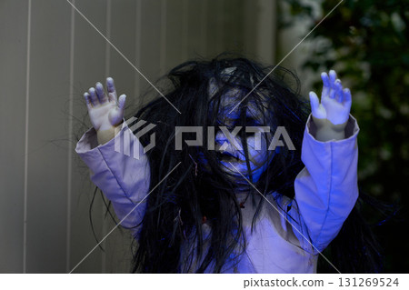 A creepy Halloween doll with white face and black messy hair raises its hands. Spooky outdoor portrait with blue light and horror mood. A creepy Halloween doll with white face and black messy hair raises its hands. Spooky outdoor portrait with blue light and horror mood. 131269524
