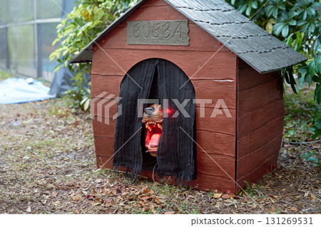 A horror dog prop snarls from a wooden doghouse with red glowing eyes. Black curtains, open jaws, and sharp fangs create a creepy outdoor Halloween scene. A horror dog prop snarls from a wooden doghouse with red glowing eyes. Black curtains, open jaws, and sharp fangs create a creepy outdoor Halloween scene. 131269531