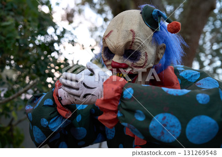 A scary clown in a green polka-dot costume holds gloved hands together. Blue hair, red nose, and yellow teeth create a spooky outdoor portrait. A scary clown in a green polka-dot costume holds gloved hands together. Blue hair, red nose, and yellow teeth create a spooky outdoor portrait. 131269548