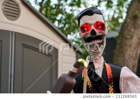 Grinning skull with red LED eyes holds a bottle beside a lighted straw. Halloween party bartender prop in vest and tie against a backyard shed. 131269563