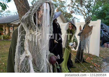 Hooded ghoul stands in the foreground while several witches gather behind a white fence near a house. Outdoor Halloween yard display with orange lights and spooky props. Hooded ghoul stands in the foreground while several witches gather behind a white fence near a house. Outdoor Halloween yard display with orange lights and spooky props. 131269584