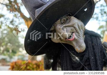 Weathered witch decoration tilts her head with a crooked smile and cracked skin. Halloween yard decor under a tree with soft background bokeh. 131269586