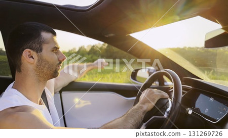 Profile of young handsome man driving a modern electric car with sunlight at background. Young male driver riding on electrical vehicle at countryside. Concept of journey and trip. Close up Profile of young handsome man driving a modern electric car with sunlight at background. Young male driver riding on electrical vehicle at countryside. Concept of journey and trip. Close up 131269726