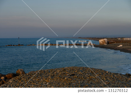 Serene coastal landscape at dusk with gentle waves lapping against rocky shore and distant silhouettes 131270312