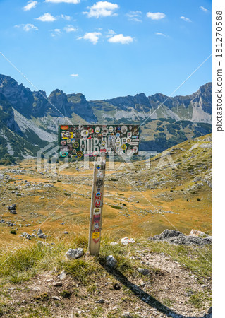 Tourist sign of Durmitor National Park in Montenegro, covered with colorful stickers from different countries, standing on a hill with scenic mountain landscape. Symbol of travel, adventure, and 131270588