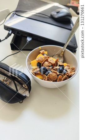 Bowl of cereal with blueberries and mango placed next to a closed laptop and glasses case on a white table Bowl of cereal with blueberries and mango placed next to a closed laptop and glasses case on a white table 131270661