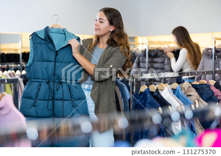Smiling female woman keeping turquoise winter waistcoat on a rack while shopping in the showroom 131275370