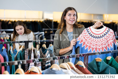 Casual young woman examining prices while holding a jumper with a graphic pattern in a clothing store Casual young woman examining prices while holding a jumper with a graphic pattern in a clothing store 131275392