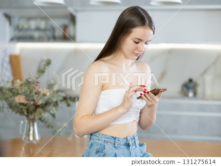 Young girl typing message on the screen of mobile phone in home kitchen Young girl typing message on the screen of mobile phone in home kitchen 131275614