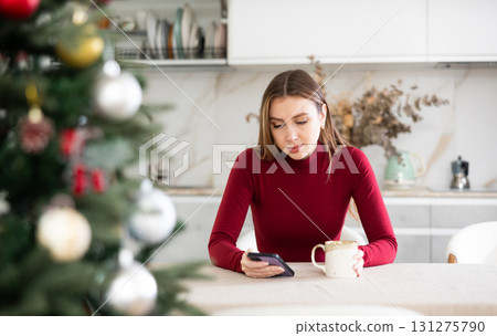 Upset lonely woman sitting at Christmas table and waiting for a call on her smartphone Upset lonely woman sitting at Christmas table and waiting for a call on her smartphone 131275790