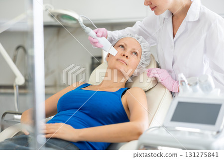 Woman lying on couch during cosmetological ultrasonic facial cleansing in clinic. Professional moisturizing and rejuvenating hardware procedures for people 131275841