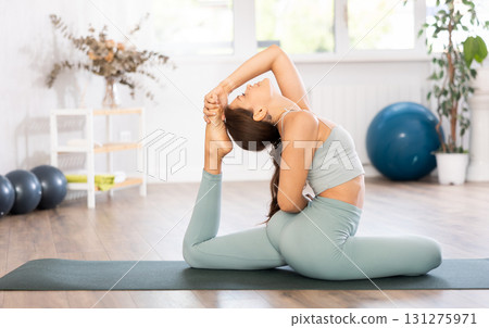 Young woman yoga trainer performing king pigeon pose on black rug 131275971