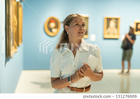 Young girl holding an information booklet looks at paintings in an art exhibition in museum 131276009