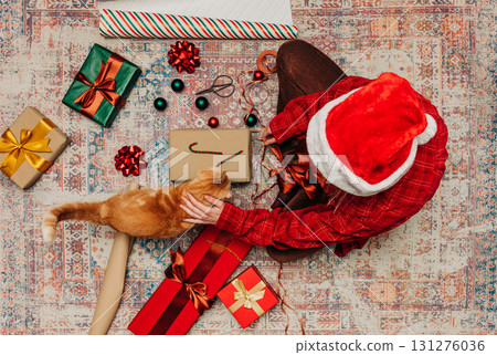 woman sits on cozy rug with boxes, busily wrapping Christmas presents, near ginger cat woman sits on cozy rug with boxes, busily wrapping Christmas presents, near ginger cat 131276036