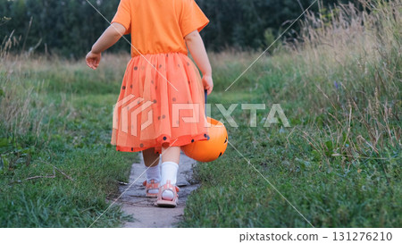 Rear view of a child girl in an orange costume walking along a path surrounded by autumn greenery holding a pumpkin shaped basket filled with candy enjoying Halloween festivities Rear view of a child girl in an orange costume walking along a path surrounded by autumn greenery holding a pumpkin shaped basket filled with candy enjoying Halloween festivities 131276210