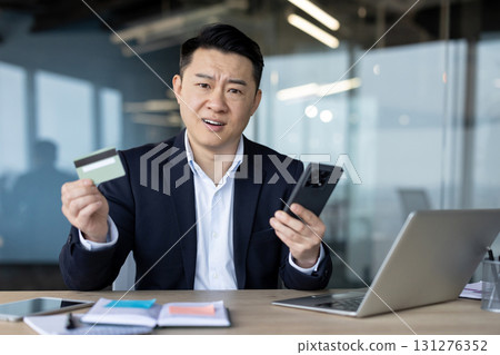 Asian businessman looking puzzled holding a credit card and a smartphone, confused by something on the screen while at his office desk. 131276352