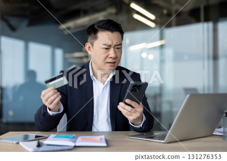Worried young Asian man sitting at his desk in the office, holding a phone and credit card, looking at the laptop screen in frustration. Worried young Asian man sitting at his desk in the office, holding a phone and credit card, looking at the laptop screen in frustration. 131276353