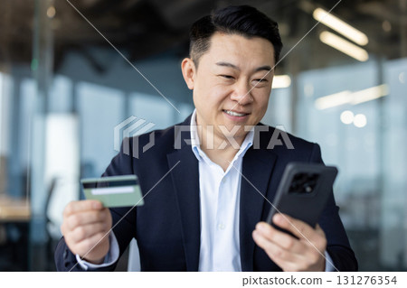 Close-up photo of a young Asian man in a suit sitting in an office, holding a credit card and using a mobile phone. Close-up photo of a young Asian man in a suit sitting in an office, holding a credit card and using a mobile phone. 131276354