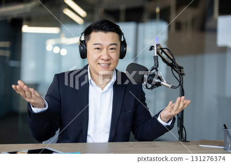 Portrait of a young Asian man sitting in the office wearing headphones in front of a desk with a microphone, talking and gesturing with his hands to the camera. 131276374