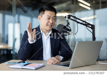 Asian young man in a suit sitting in the office in front of a desk with a microphone and talking, on a video call on a laptop. 131276387