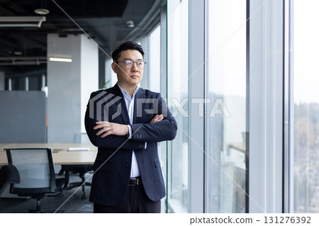 Asian young male businessman standing in the office near the window wearing a suit and glasses, arms crossed on his chest and looking to the side. Asian young male businessman standing in the office near the window wearing a suit and glasses, arms crossed on his chest and looking to the side. 131276392