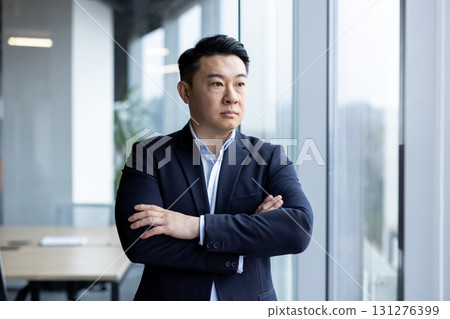Asian young successful and serious businessman standing in a suit in the office, arms crossed on his chest, looking out the window. Asian young successful and serious businessman standing in a suit in the office, arms crossed on his chest, looking out the window. 131276399
