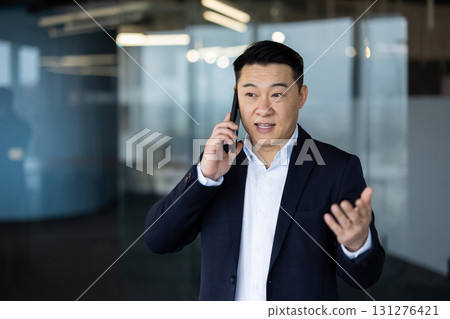A young Asian male businessman in a business suit is standing inside an office center, talking on a mobile phone and gesturing with his hands. 131276421